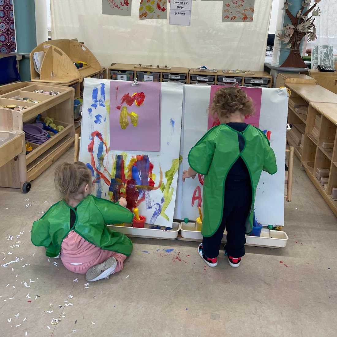Two children in aprons painting side by side at a double easel